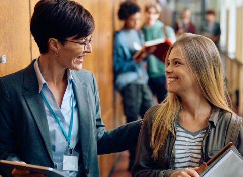 Happy teenage girl talking to her teacher in high school hallway.