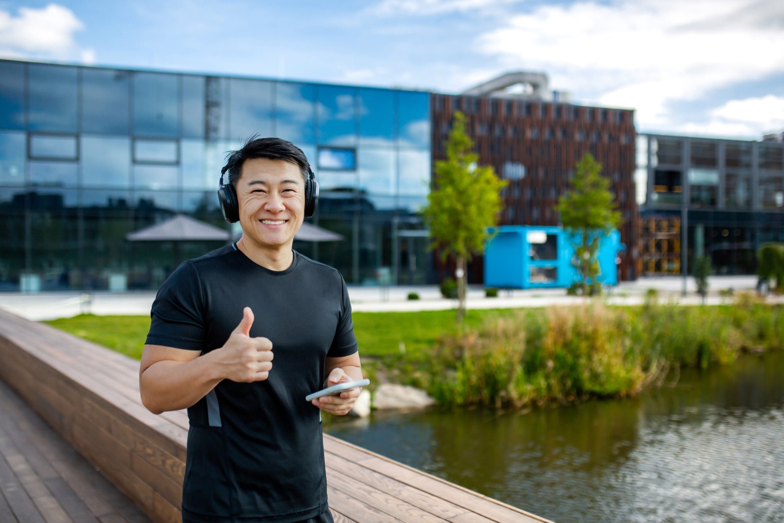 Portrait of a young Asian sportsman standing outside wearing headphones, holding a mobile phone, smiling at the camera and showing super